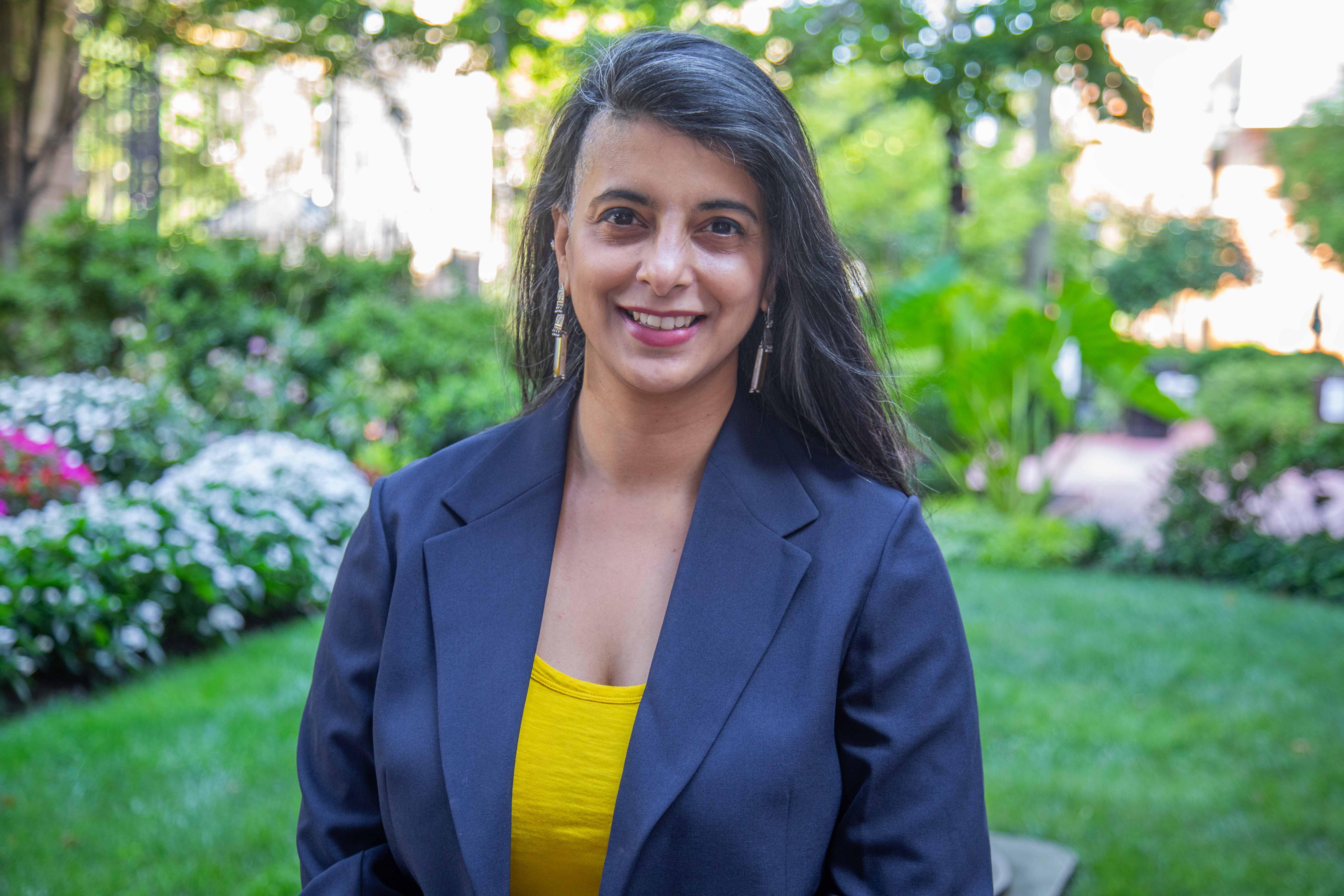 Shayoni Mitra headshot. She is wearing a yellow shirt and black blazer. Campus greenery is behind her