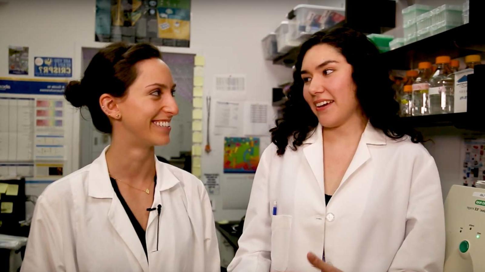 Two young women in lab coats speaking in a science lab