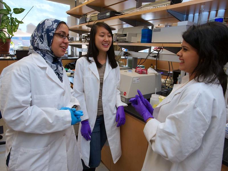3 young women of color in white lab coats discuss something in a lab
